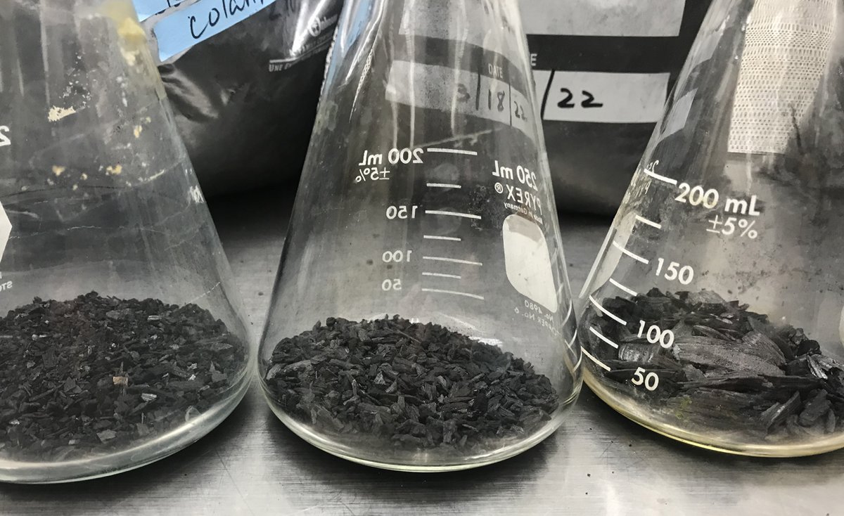 Biochar from hardwood materials in three glass flasks on a metal lab bench.